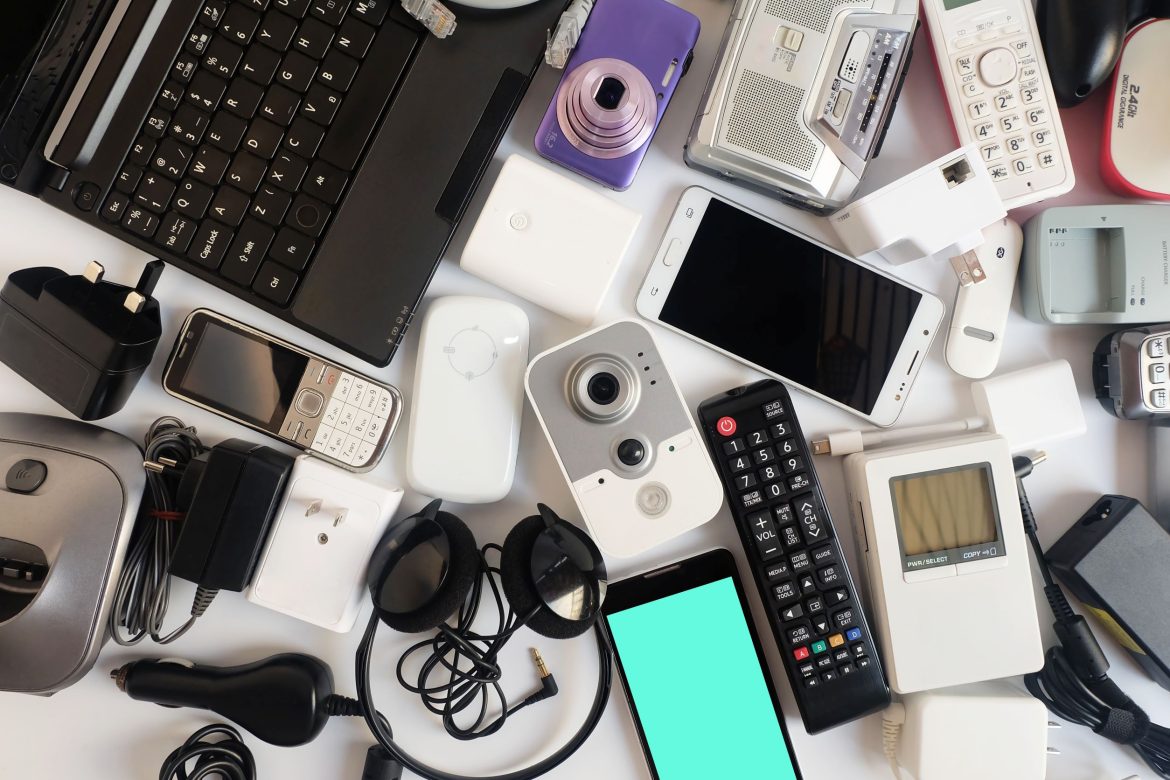 Selection of unwanted electronic items on a table, including laptop, chargers, mobile phones, plugs, camera, headphones, remote control.