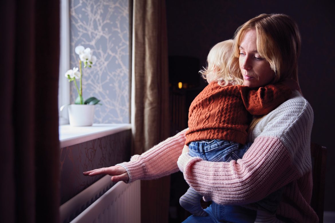 A woman stands at a window cradling a toddler checking to see if the radiator is warm.