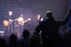 An audience member photographed from the back takes a picture of Michael Head and The Red Elastic Band, on stage at Knowsley Leisure and Culture Park
