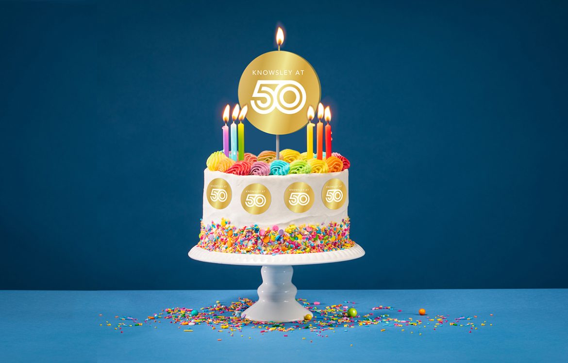 Birthday cake with rainbow icing, colorful Sprinkles and lit candles over a blue background. Topper says "Knowsley at 50"